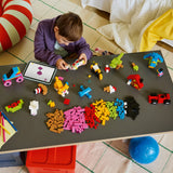 Niño construyendo con LEGO Classic 11045 sobre mesa de juego vista cenital, con tableta de instrucciones LEGO Builder y ladrillos de colores: rosa, naranja, amarillo, azul y modelos ya montados