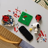 Vista cenital de mesa con piezas LEGO Ideas 21368 Peanuts a media construcción: base verde de la caseta con etiqueta SNOOPY, cabeza de Snoopy montada y bowl de ladrillos rojos preparados