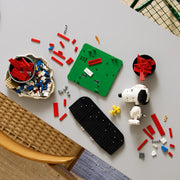 Vista cenital de mesa con piezas LEGO Ideas 21368 Peanuts a media construcción: base verde de la caseta con etiqueta SNOOPY, cabeza de Snoopy montada y bowl de ladrillos rojos preparados