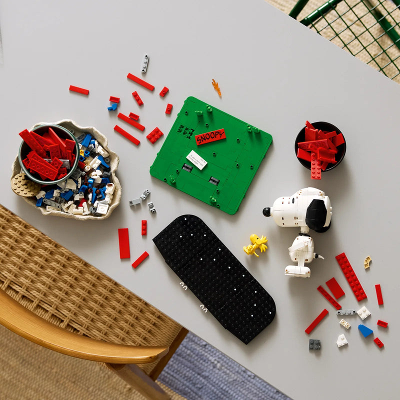 Vista cenital de mesa con piezas LEGO Ideas 21368 Peanuts a media construcción: base verde de la caseta con etiqueta SNOOPY, cabeza de Snoopy montada y bowl de ladrillos rojos preparados