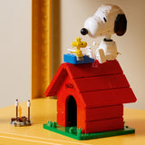 LEGO Ideas 21368 Snoopy en su caseta de Peanuts expuesto sobre superficie amarilla junto a un mini-set LEGO Camping con hoguera - decoración coleccionista adulta