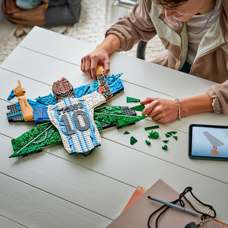 Adulto construyendo el mosaico LEGO Editions 43018 Messi sobre mesa con instrucciones en tableta - set premium 14+ para fans del fútbol