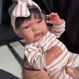 Baby Toneta 60146 de Muñecas Antonio Juan en brazos, foto real con luz natural: pijama de rayas crema y rosa y diadema de lazo