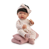 Baby Toneta de Antonio Juan con pijama de rayas crema y rosa empolvado, diadema con lazo y chupete, sobre su manta palo de rosa