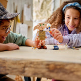 Niño con gorra y niña con turbante azul sonriendo y jugando con la figura LEGO Harry Potter 76469 Dobby el Elfo Libre con su libro y estrellas mágicas sobre mesa de madera