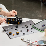 Manos adultas construyendo LEGO Speed Champions 77262 Ken Block Mustang Hoonicorn sobre cartel HOONIGAN, con pegatinas, ruedas doradas y tableta LEGO Builder