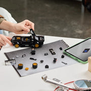 Manos adultas construyendo LEGO Speed Champions 77262 Ken Block Mustang Hoonicorn sobre cartel HOONIGAN, con pegatinas, ruedas doradas y tableta LEGO Builder