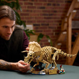 Hombre adulto con tatuajes sonriendo mientras articula la cabeza del esqueleto LEGO Jurassic World 77985 Triceratops sobre mesa con pared de ladrillo - set premium 18+ para fans