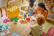 Familia construyendo con la caja de ladrillos creativos LEGO Classic 10698 sobre la mesa, con el cubo amarillo de almacenamiento