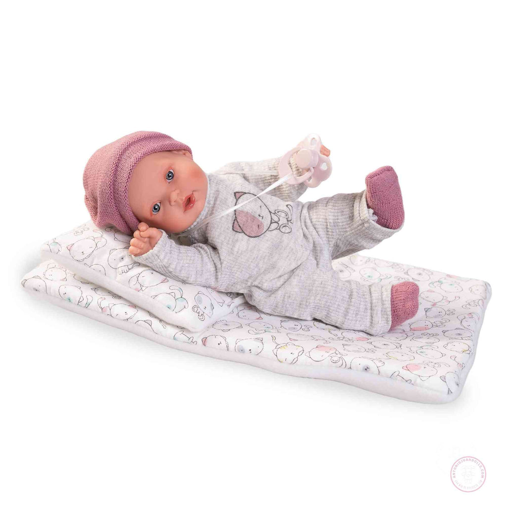 Muñeca Kika Lagrimitas Antonio Juan 11115 sobre su camita con gorrito rosa, pijama gris con gatito y chupete — vista lateral