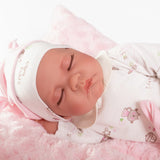 Primer plano de la cara de la muñeca Luna de Antonio Juan dormida, con gorro y pijama blanco sobre cojín rosa