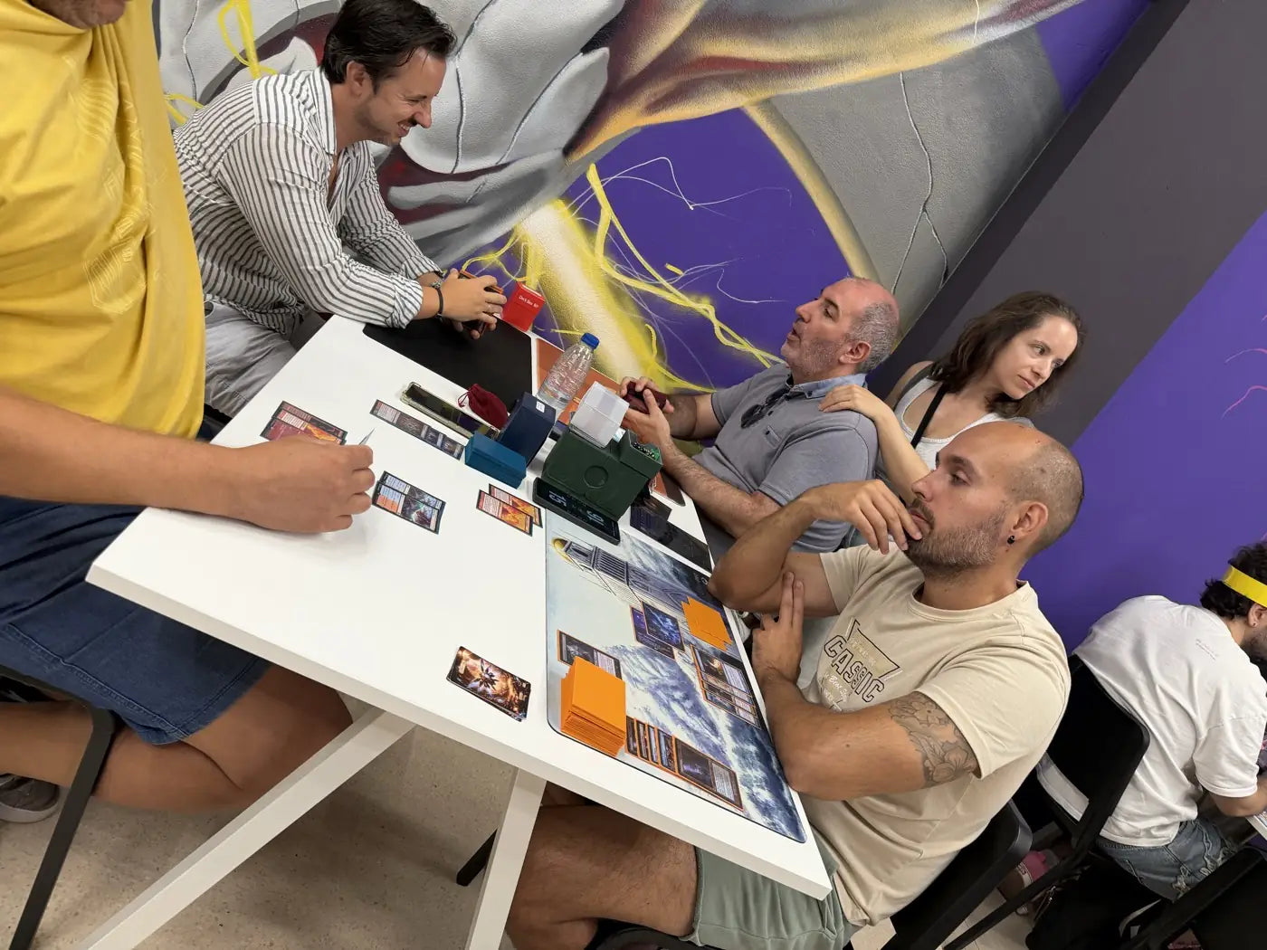 Una mesa de la ronda suiza, con cuatro jugadores y el mural de la tienda detrás